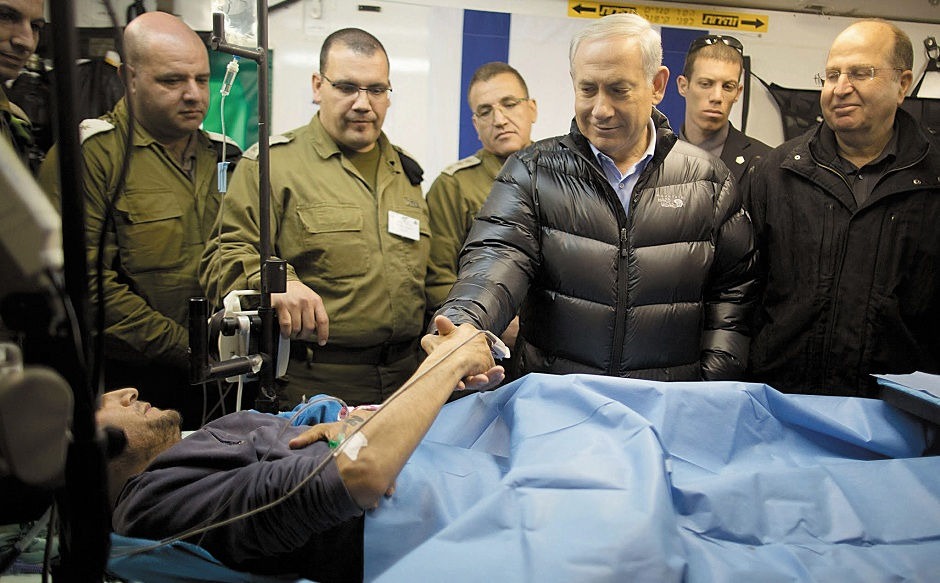 ISRAELS LEDNING HÄLSAR PÅ ”SYRISKA REBELLER” på ett israeliskt fältsjukhus den 18 februari 2014. I en video syns premiärminister Benjamin Netanyahu till och med skaka hand med en av ”rebel­lerna”. Med honom var flera ur Israels politiska och militära toppskikt, däribland dåvarande försvarsministern Moshe Ya’alon (t.h.). Dåvarande generalstabschefen Benny Gants (utanför bild) kan ses i videon. Han bär en automatkarbin och ser synbart besvärad ut. När bilderna spreds över världen tystnade snabbt anklagelser om att tidigare rapporter från 2013 om israelisk vård av ”syriska rebeller” var ”antisemitiska lögner”. Wall Street Journal (WSJ) skrev i samband med besöket att en israelisk militär tjänsteman berättat att ”Vi frågar inte vilka de [vårdtagande syriska rebellerna] är, vi gör ingen kontroll”. Han sade vidare att ”När behandlingen är klar tar vi dem tillbaka till gränsen där de går vidare [in i Syrien]”. Tjänstemannen sade även till WSJ att det finns en ”tyst överenskommelse” mellan israeliska styrkor och de så kallade syriska rebellerna, som flera medier inklusive MintPress News utan omskrivning beskriver som ”al-Qaida krigare”. Israels officiella försäkringar att man är opartisk i den syriska konflikten klingar därför allt mer ihåligt. Stillbild: ”The Prime Minister of Israel” YouTube kanal